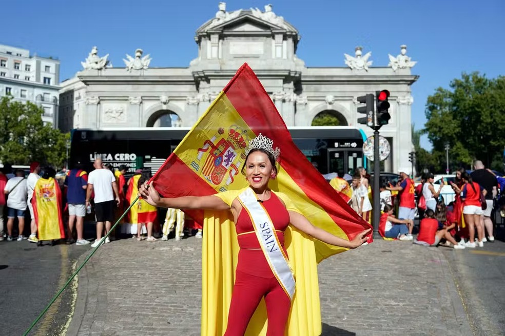 Uma mulher posa segurando uma bandeira da Espanha enquanto torcedores aguardam a passagem do desfile dos jogadores da seleção, um dia após a conquista do segundo título da Copa do Mundo com a vitória por 1 a 0 sobre a Argentina, na Puerta de Alcalá, em Madri, em 20 de julho de 2026. Os campeões mundiais retornaram à capital espanhola para uma recepção de heróis, onde cerca de um milhão de torcedores eram esperados para saudá-los após uma noite de intensas comemorações por todo o país — Foto: Paul Hanna / AFP